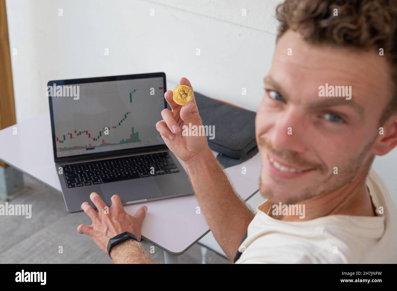 Young smiling trader is making money from cryptocurrency trading, he is holding a bitcoin coin and the candlestick chart is green and positive Stock Photo