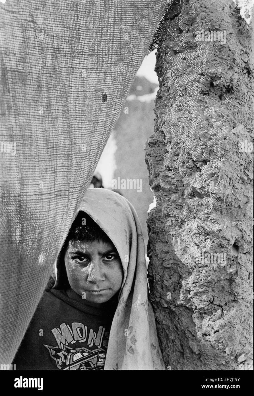 A young Afghan girl, looks through a jute curtain, standing at the door of her mud house, in Mohammad Khel Refugee Camp, in Balauchistan Province, in Stock Photo
