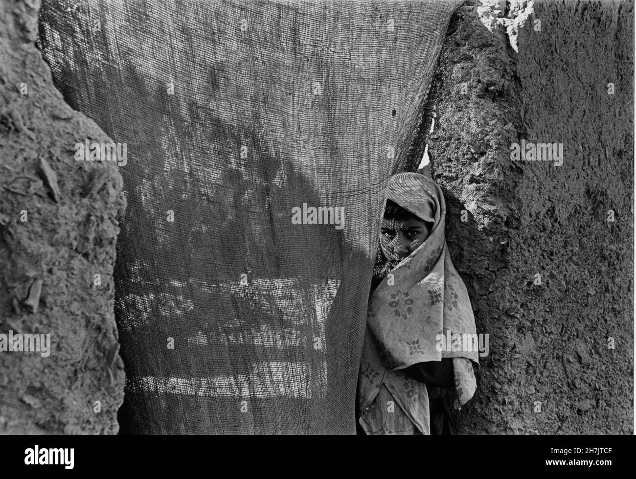 A young Afghan girl, looks through a jute curtain, standing at the door of her mud house, in Mohammad Khel Refugee Camp, in Balauchistan Province, in Stock Photo