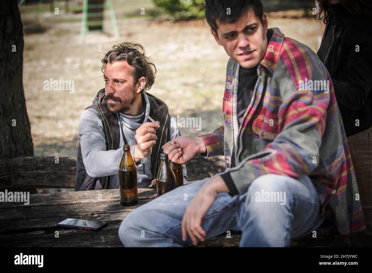 Young friends keep watch, look around and try to hide while gives from hand to hand each other ganja joint in public park outdoor. Smoking cannabis ma Stock Photo