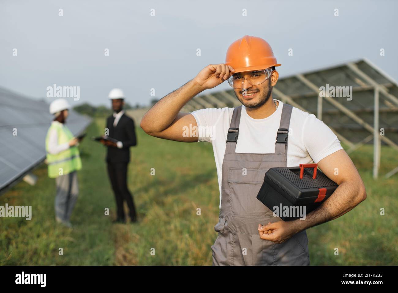 Indian technician touching his orange helmet and smiling on camera while two engineers talking behind. Multiracial people working on big station with solar panels. Stock Photo