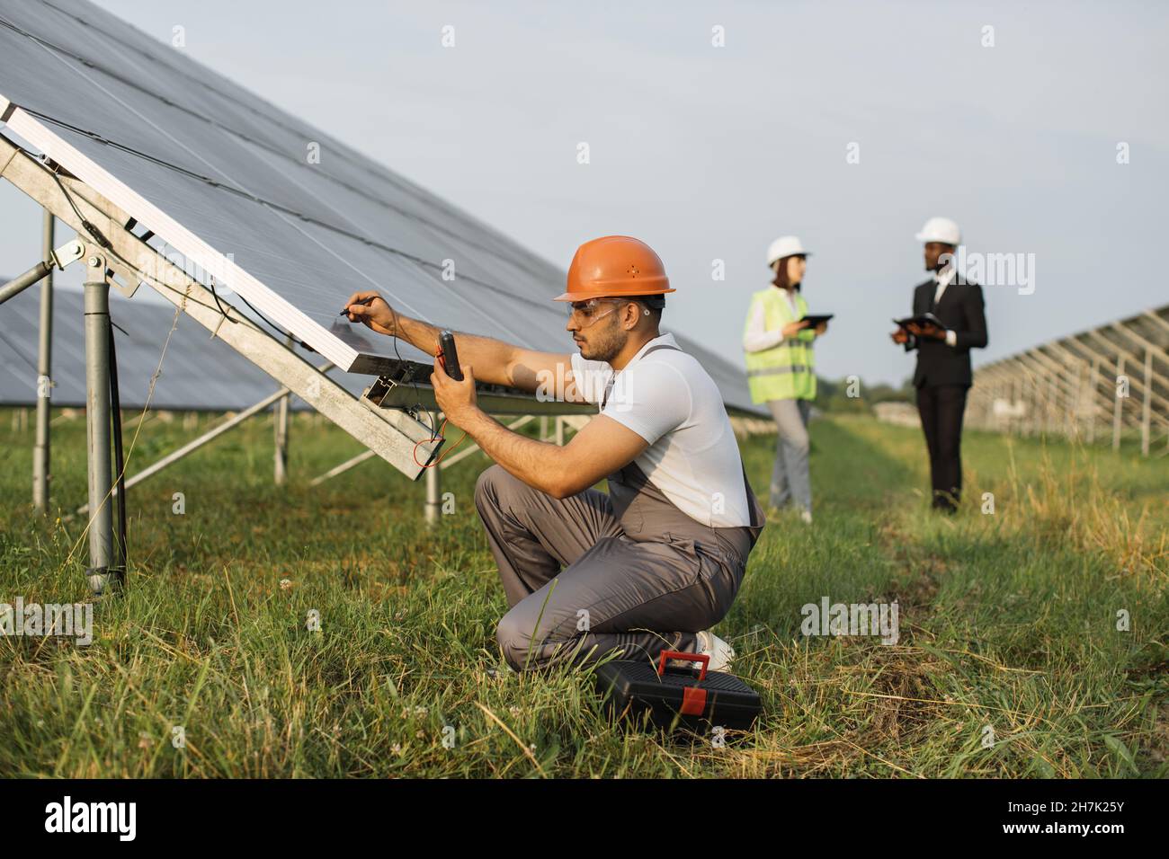 Energy technician in helmet and uniform using multimeter for measuring voltage of solar panels outdoors. Group of engineers with tablet and clipboard standing on background. Stock Photo