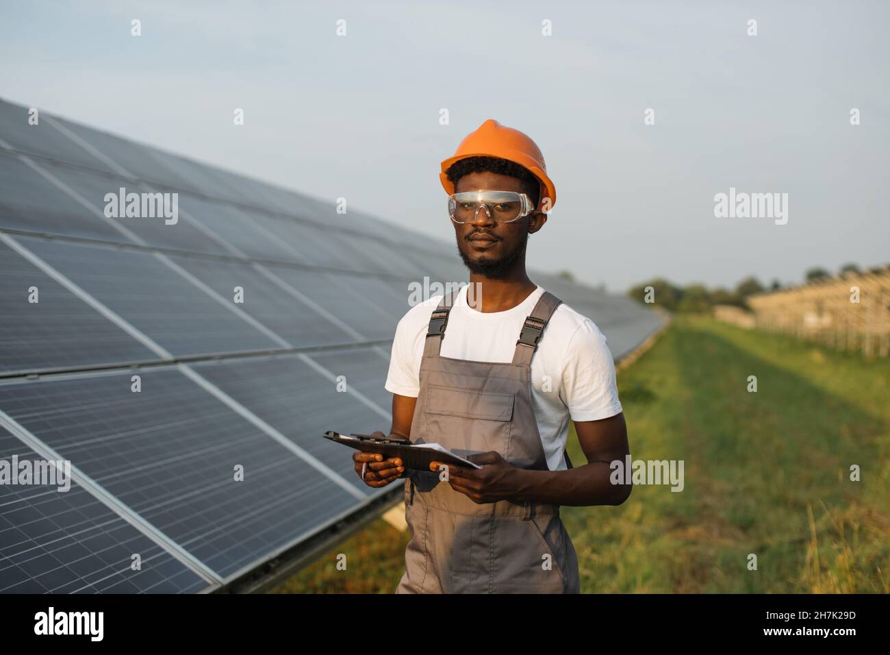 Portrait of african american man in safety glasses and orange helmet holding clipboard while standing on field with solar panels. Concept service work and alternative energy. Stock Photo