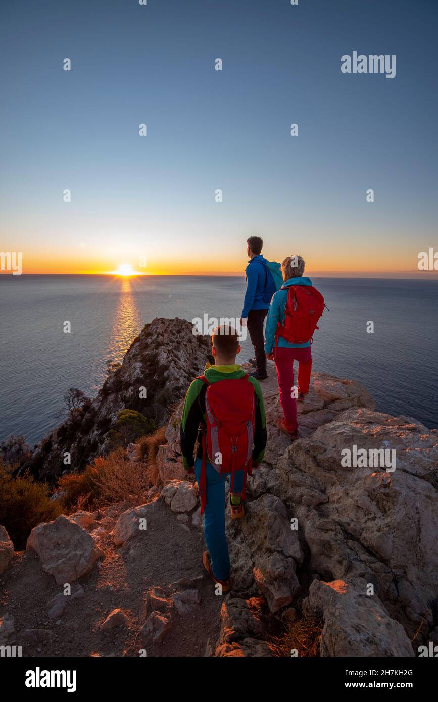 Three hikers stand on pinnacle above the mediterranean sea and hills and look out, Calpe, Alicante province, Spain Stock Photo