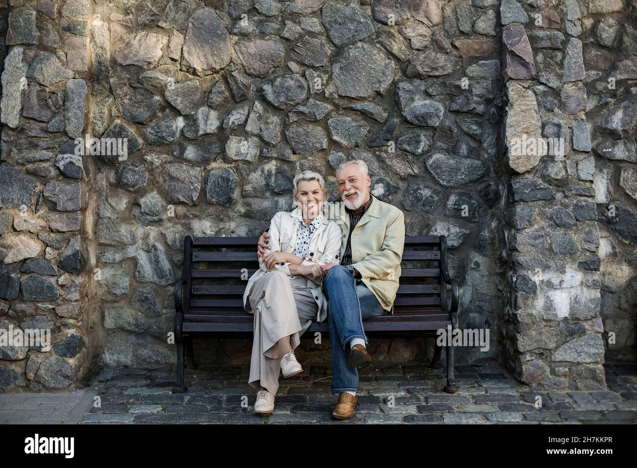 Smiling couple sitting with legs crossed at knee in front of stone wall Stock Photo