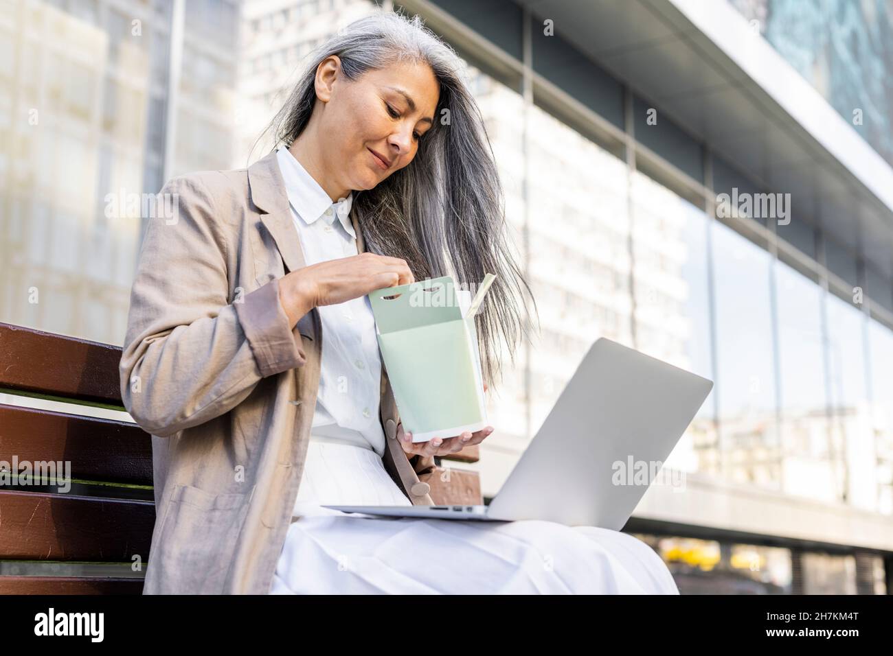 Woman with laptop eating meal while sitting on bench Stock Photo