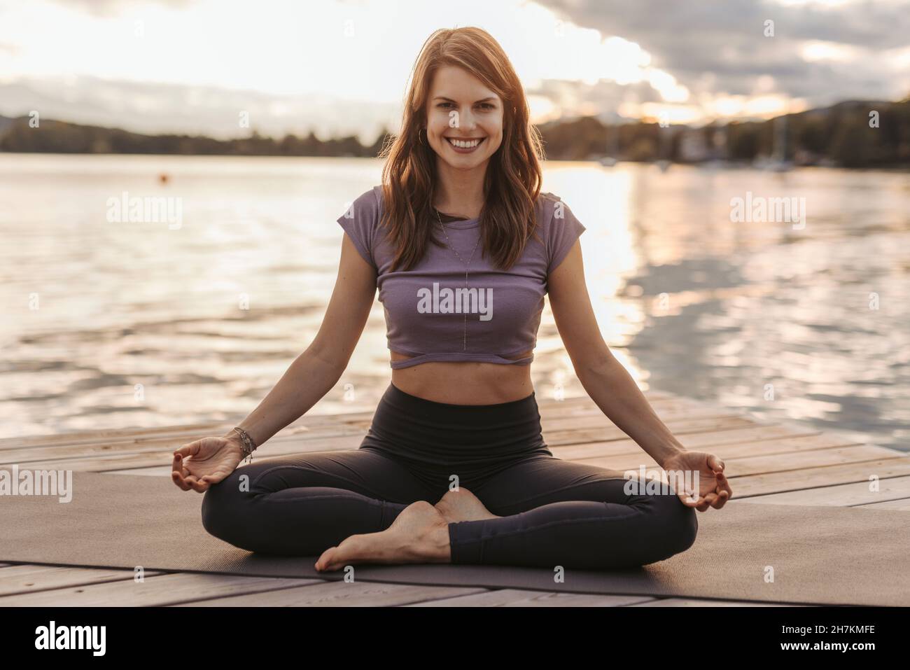 Smiling woman practicing lotus position while sitting on exercise mat by lake during sunset Stock Photo