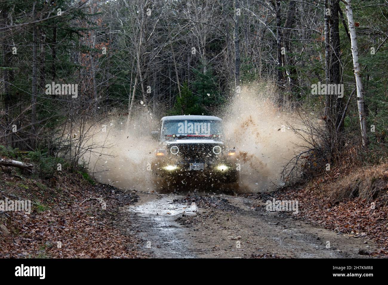 A Jeep Wrangler driving through a frozen mud puddle sending water and ice through the air on a logging road in the Adirondack Mountains, NY Stock Photo