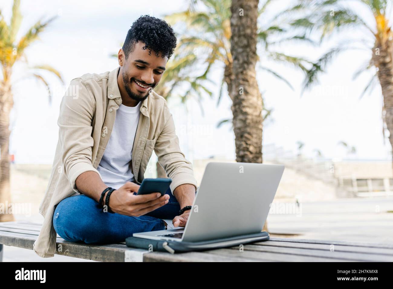 Smiling man with laptop using mobile phone on bench Stock Photo