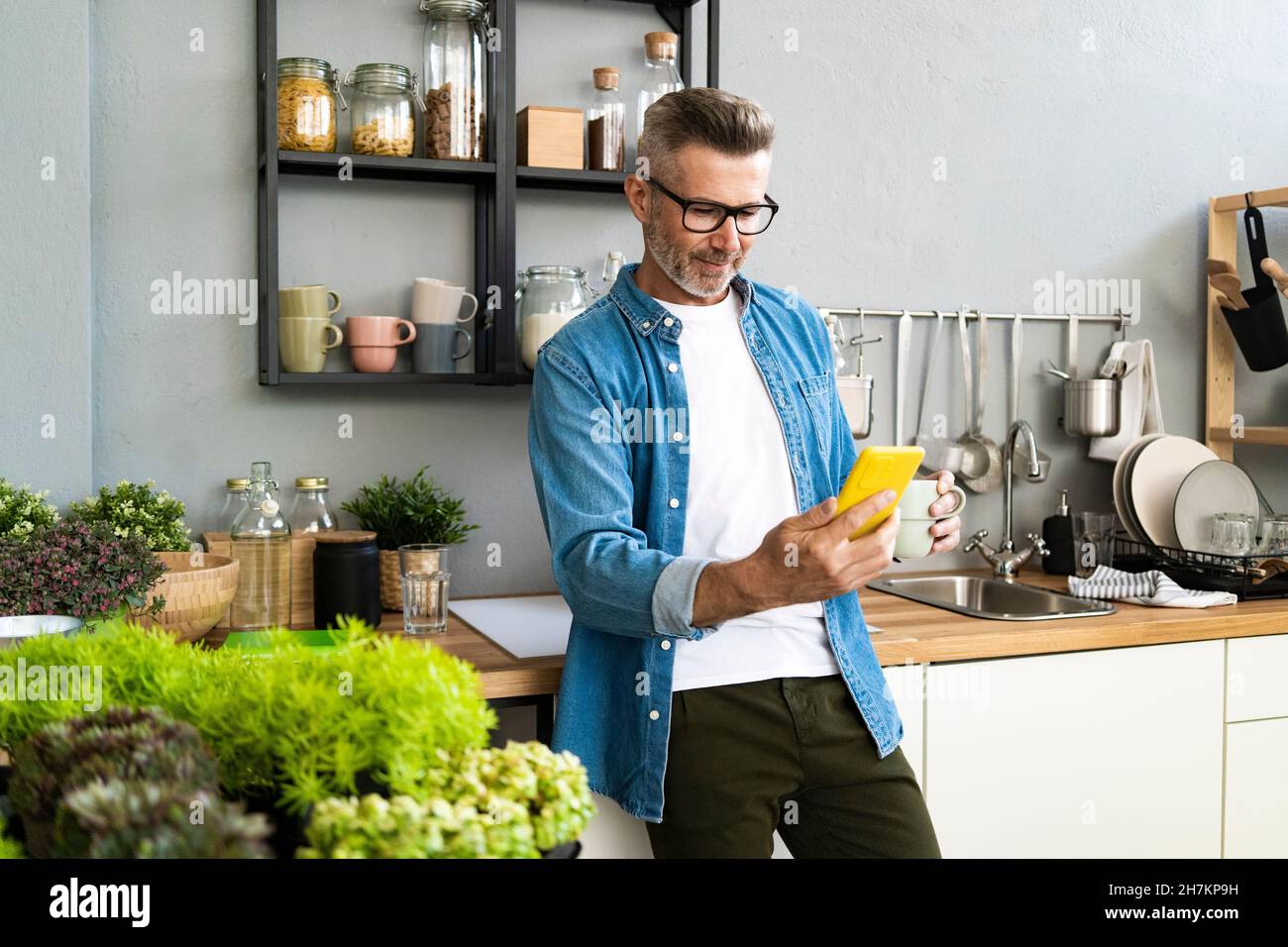 Man with coffee cup using mobile phone in kitchen at home Stock Photo