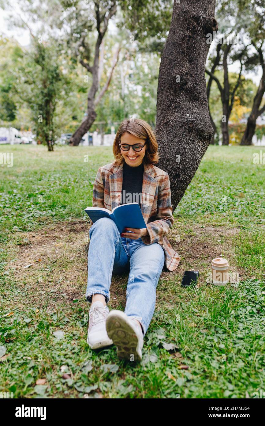Smiling freelancer reading book in front of tree in park Stock Photo