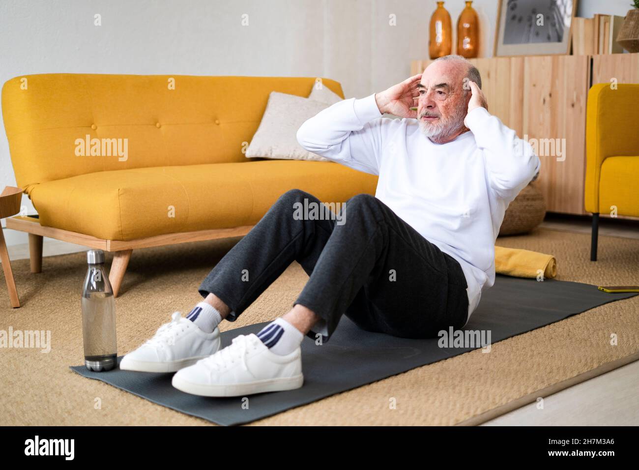Smiling senior man with hands behind head exercising on mat at home Stock Photo