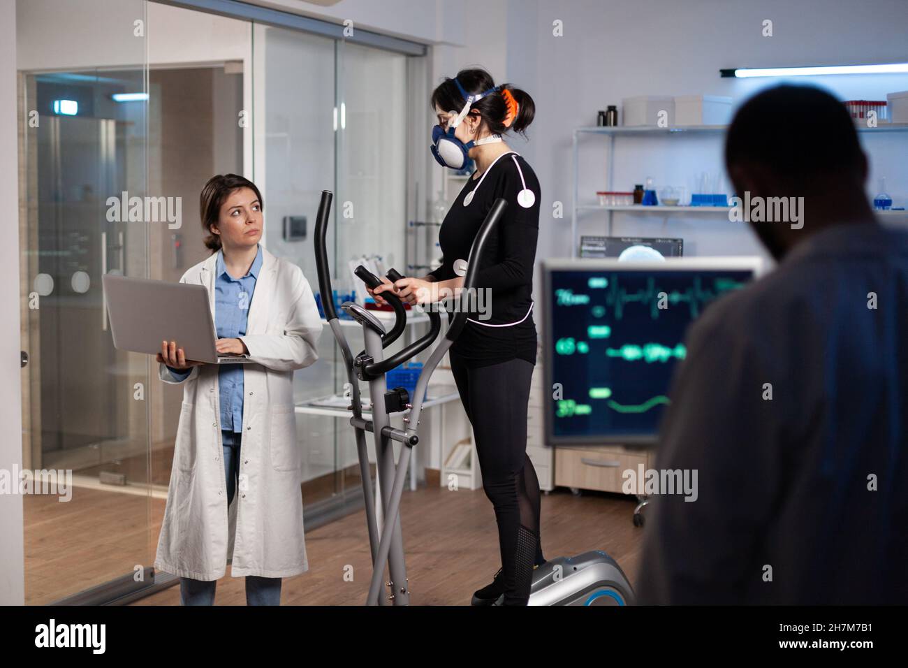 Specialist fitness researcher monitoring endurance controling EGK of sportwoman with mask running on cross trainer. Physician man analyzing heart rate in medical modern laboratory. Medicine service Stock Photo