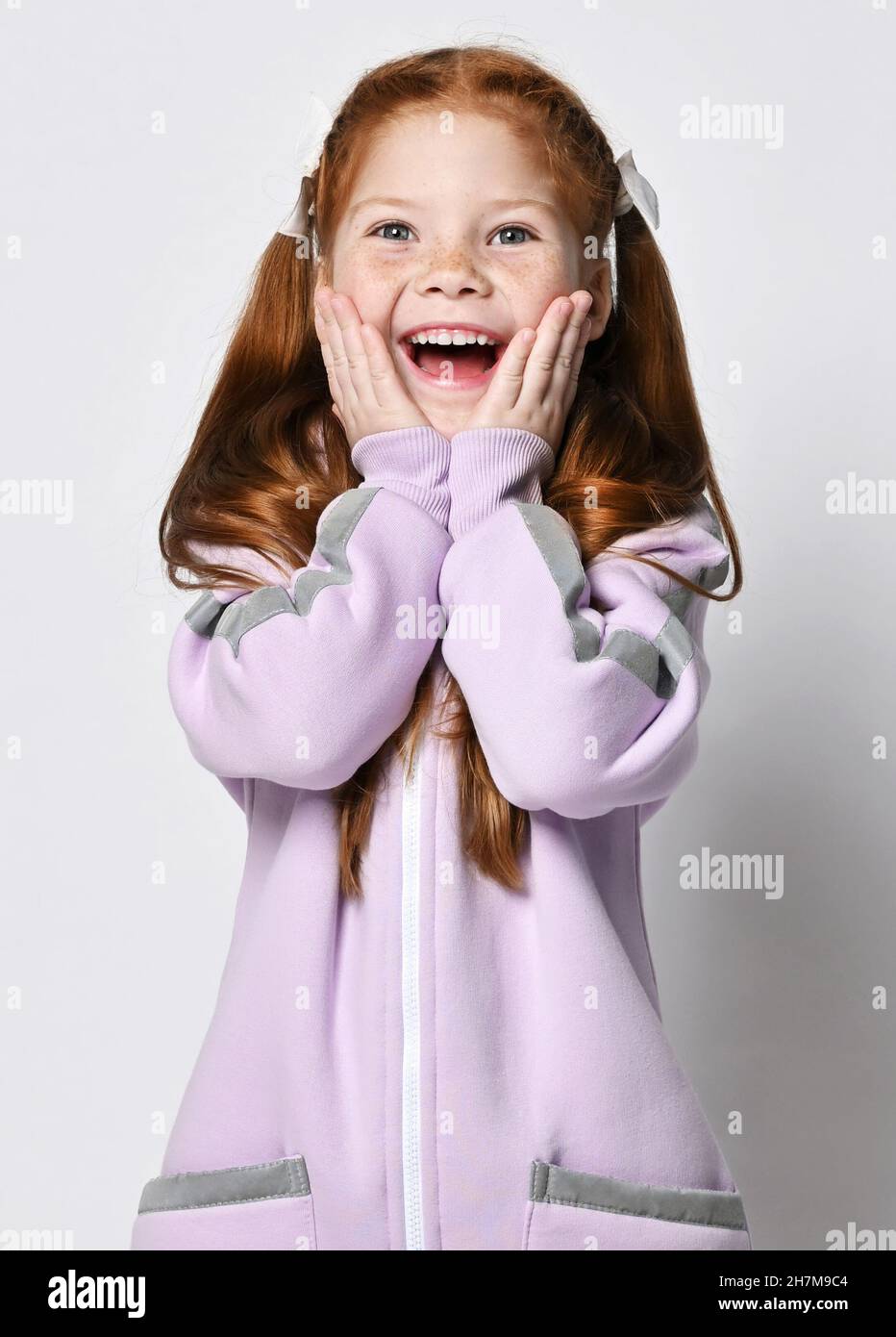 Portrait of happy, amazed kid girl in pink jumpsuit or hoodie with zipper holding her mouth open and hands at cheeks Stock Photo