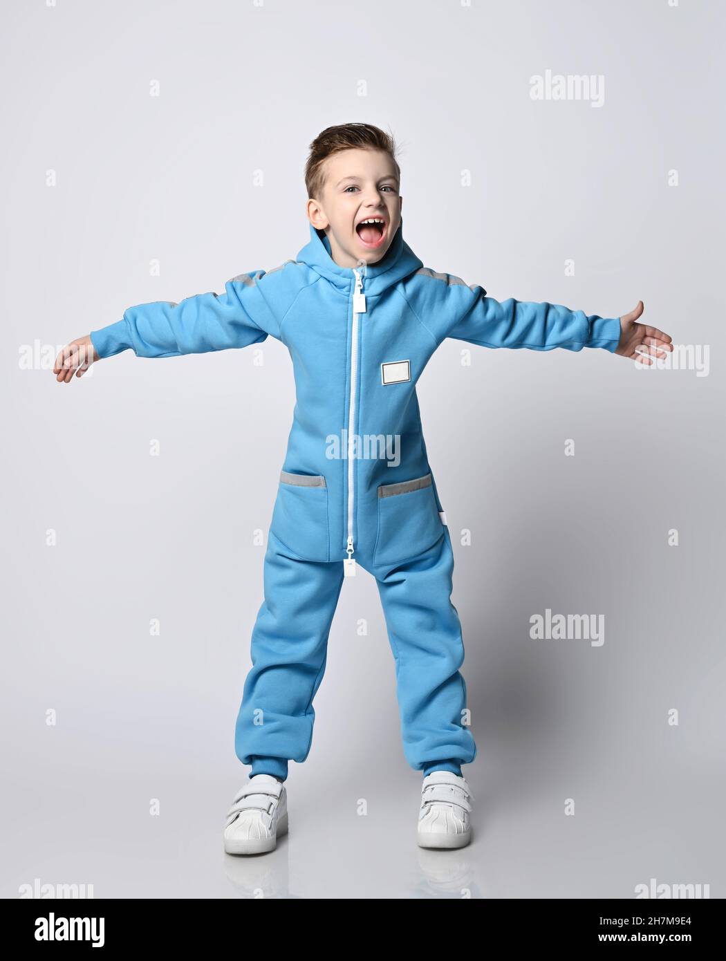 Frolic kid boy in blue jumpsuit with zipper and pockets stands holding arms wide apart and scream loudly Stock Photo