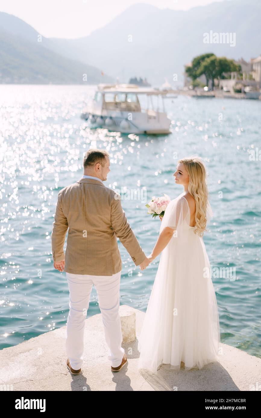 The groom and the bride with a bouquet stand holding hands on the pier ...