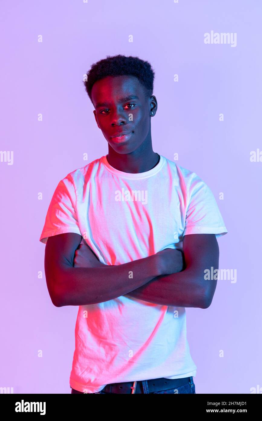 Self assured young African American male model with dark hair in white t shirt standing against purple background with crossed arms and looking at cam Stock Photo