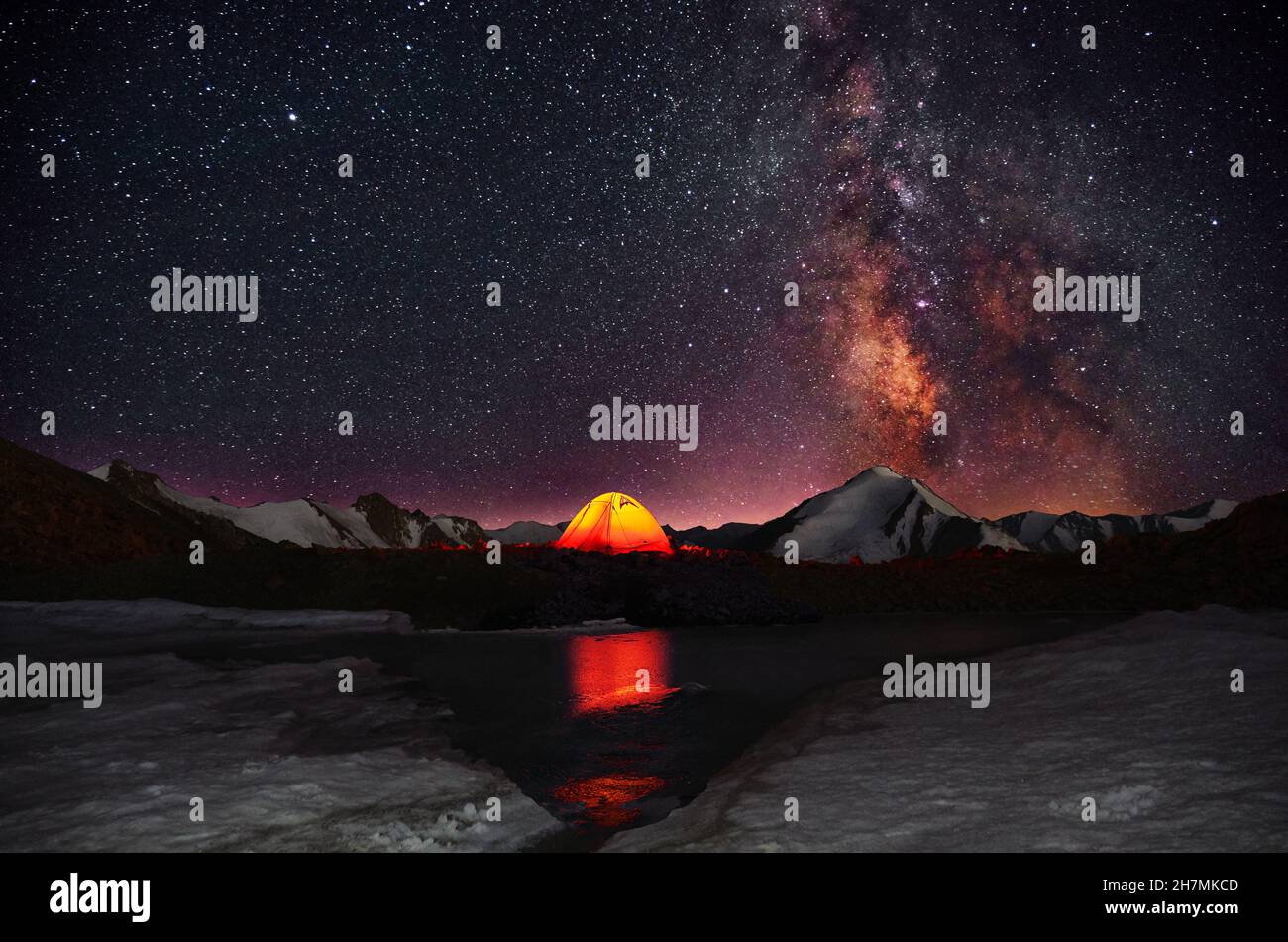 Brightly glowing orange tent set up in a romantic place on the shore of a glacial lake on the background of snow-capped mountain peaks and a starry sk Stock Photo