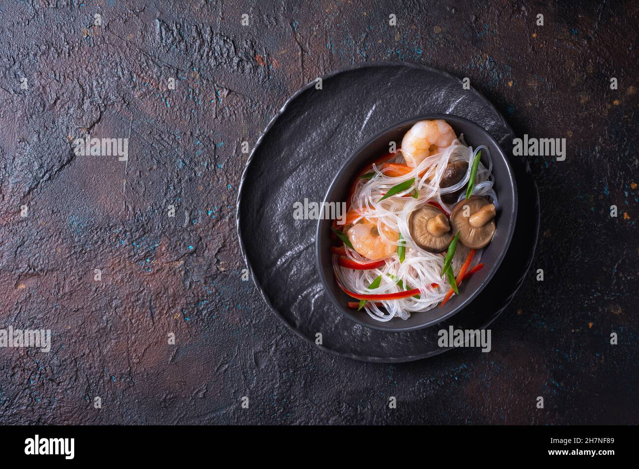 Top view of bowl with japanese bean or glass noodles, tasty shrimps, shiitake mushrooms, green onion and red pepper on dark concrete background Stock Photo