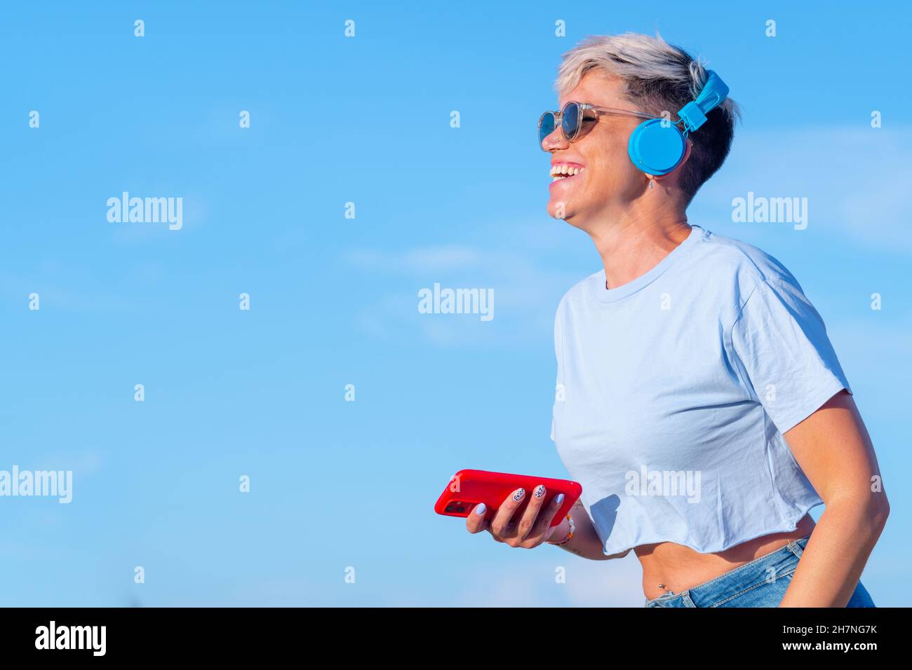 pretty young woman with short blond hair, sunglasses and short t-shirt showing her belly button has fun and smiles listening to music with wireless he Stock Photo