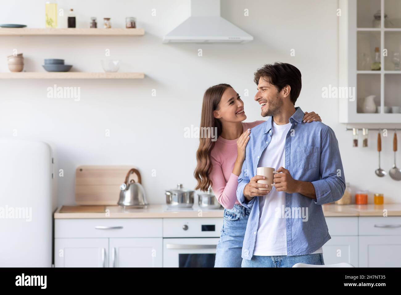 Cheerful millennial european wife hugs husband with cup of hot drink in minimalist kitchen interior Stock Photo