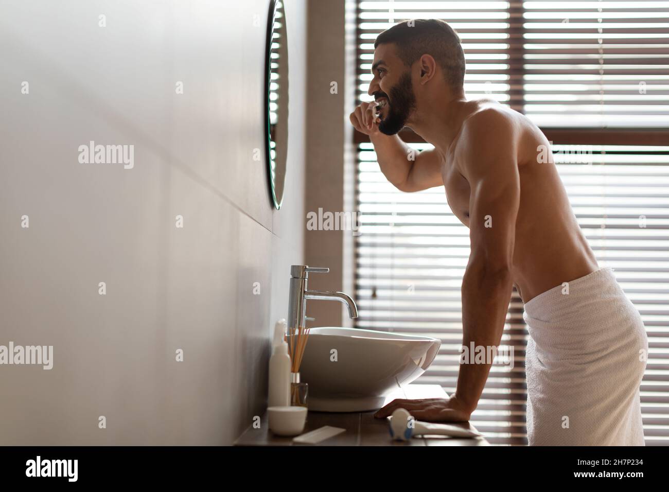 Handsome young Arab man using toothbrush in bathroom, profile view Stock Photo