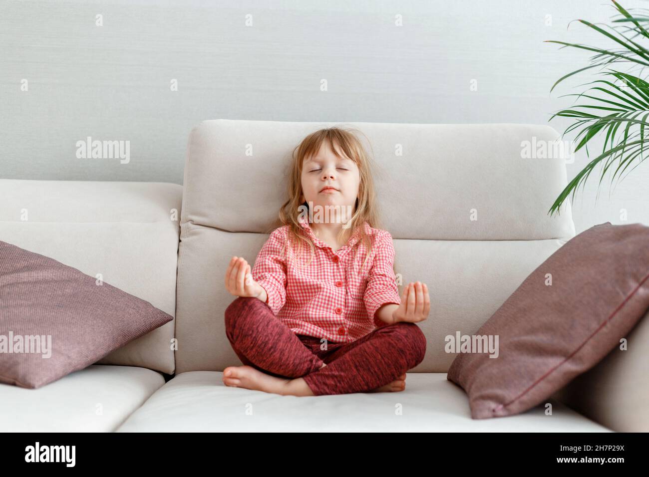 Little caucasian 3 year girl meditates on sofa while practicing yoga. Calm Kid Girl Daughter in pajamas with close eyes meditates in morning in home Stock Photo