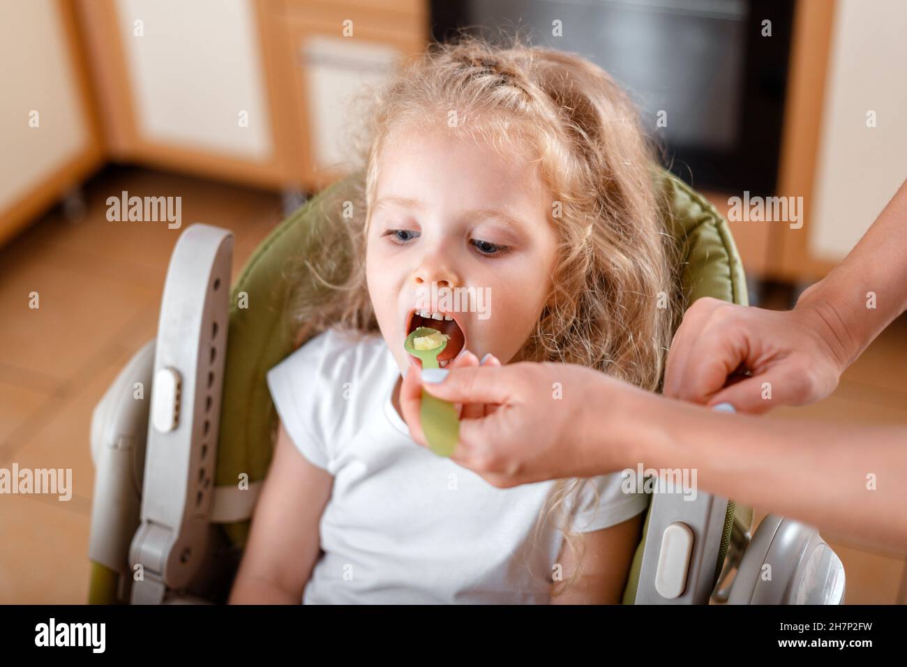 Beautiful little girl blonde eating from spoon eats porridge. Female mother's hands are feeding daughter baby child Caucasian girl in High Chair at Stock Photo