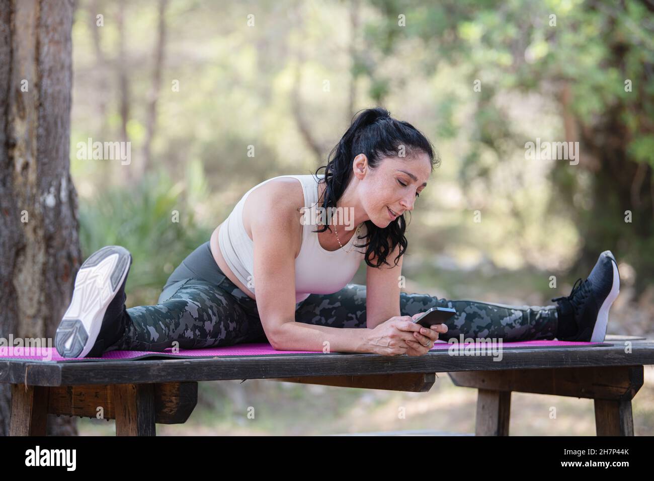 Caucasian adult woman sitting with open legs flexibility posing on picnic table using her phone Stock Photo