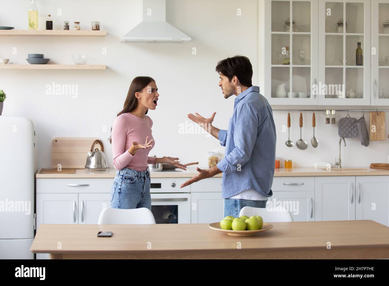 Angry excited caucasian millennial husband and wife quarreling, yelling at each other and gesturing in kitchen Stock Photo
