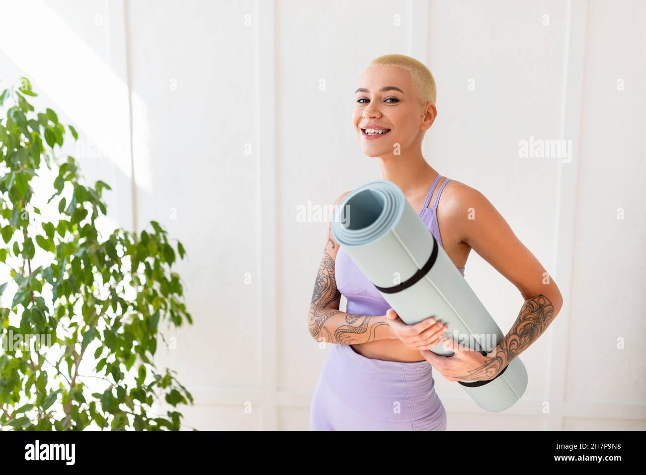 Portrait of positive woman holding yoga mat and smiling at camera while standing against white wall, ready for training Stock Photo