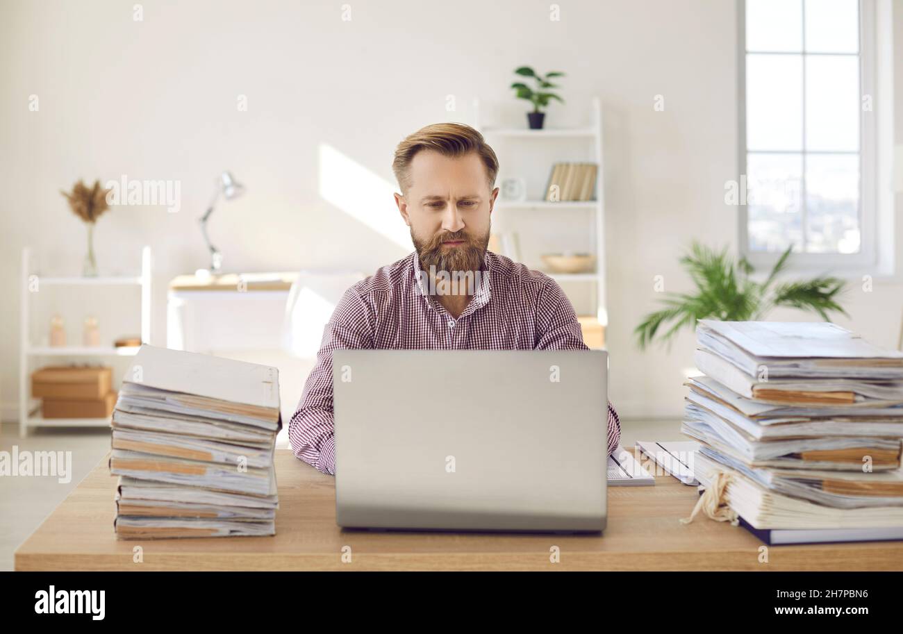 Portrait of serious and focused manager working on laptop among piles of documents. Stock Photo