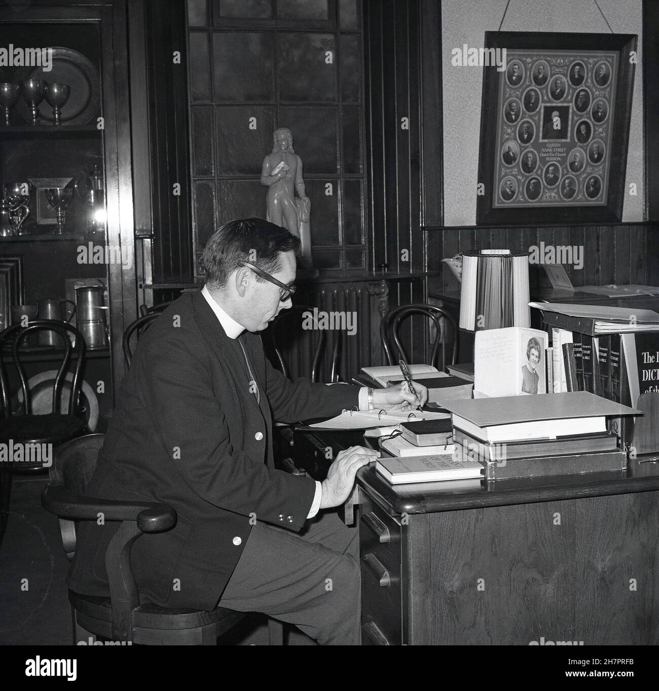 1965, historical, inside a room at a church, a young male minister of the Church of Scotland, sitting at his desk, ink-pen in hand writting notes, with various reference books to hand, including Dictionary of the Bible and Minister's Prayer Book, Fife, Scotland, UK. Stock Photo