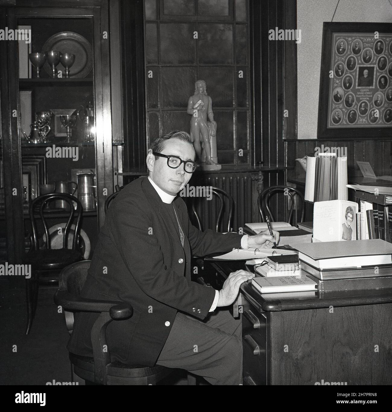 1965, historical, inside a room at a church, a young male minister of the Church of Scotland, sitting at his desk, ink-pen in hand writting notes, with various reference books to hand, including Dictionary of the Bible and Minister's Prayer Book, Fife, Scotland, UK. Stock Photo