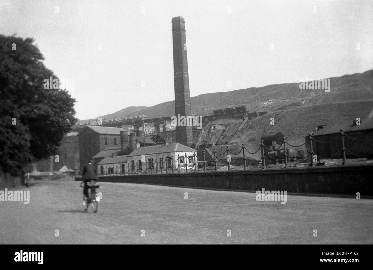1935, historical, a young man cycling past a colliery in the valley at Ebbw Vale, Wales, UK. From the1800s, Ebbw Vale had developed into a major centre for the production of iron and steel, powered by the coal from tthe numerous mines in the area. In the picture we can see the mine stack or chimney which was attached to the engine house and which interestingly is a square construction as opposed to the normal round. Stock Photo