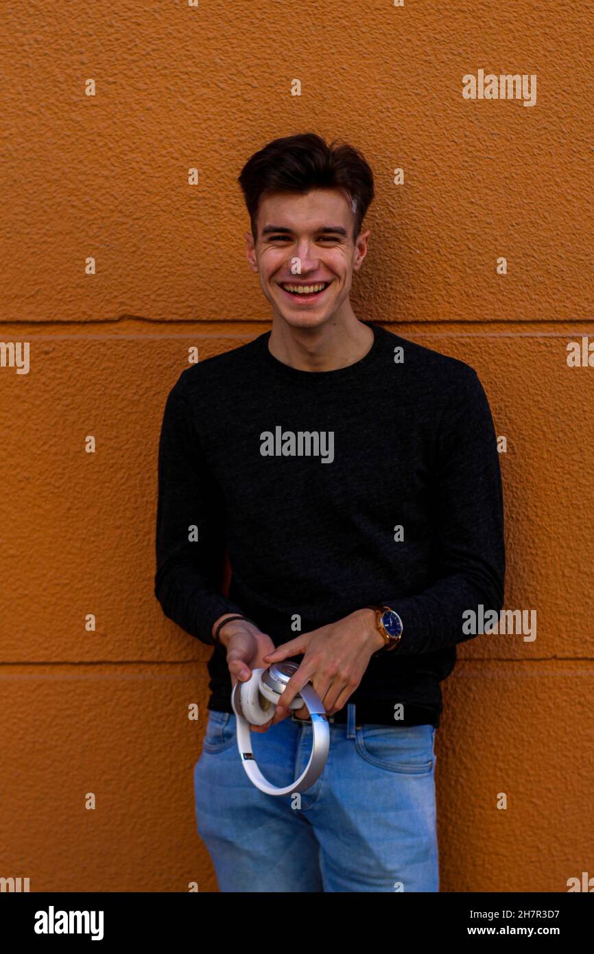 Smiling handsome Spanish male holding a headphone leaning on a wall