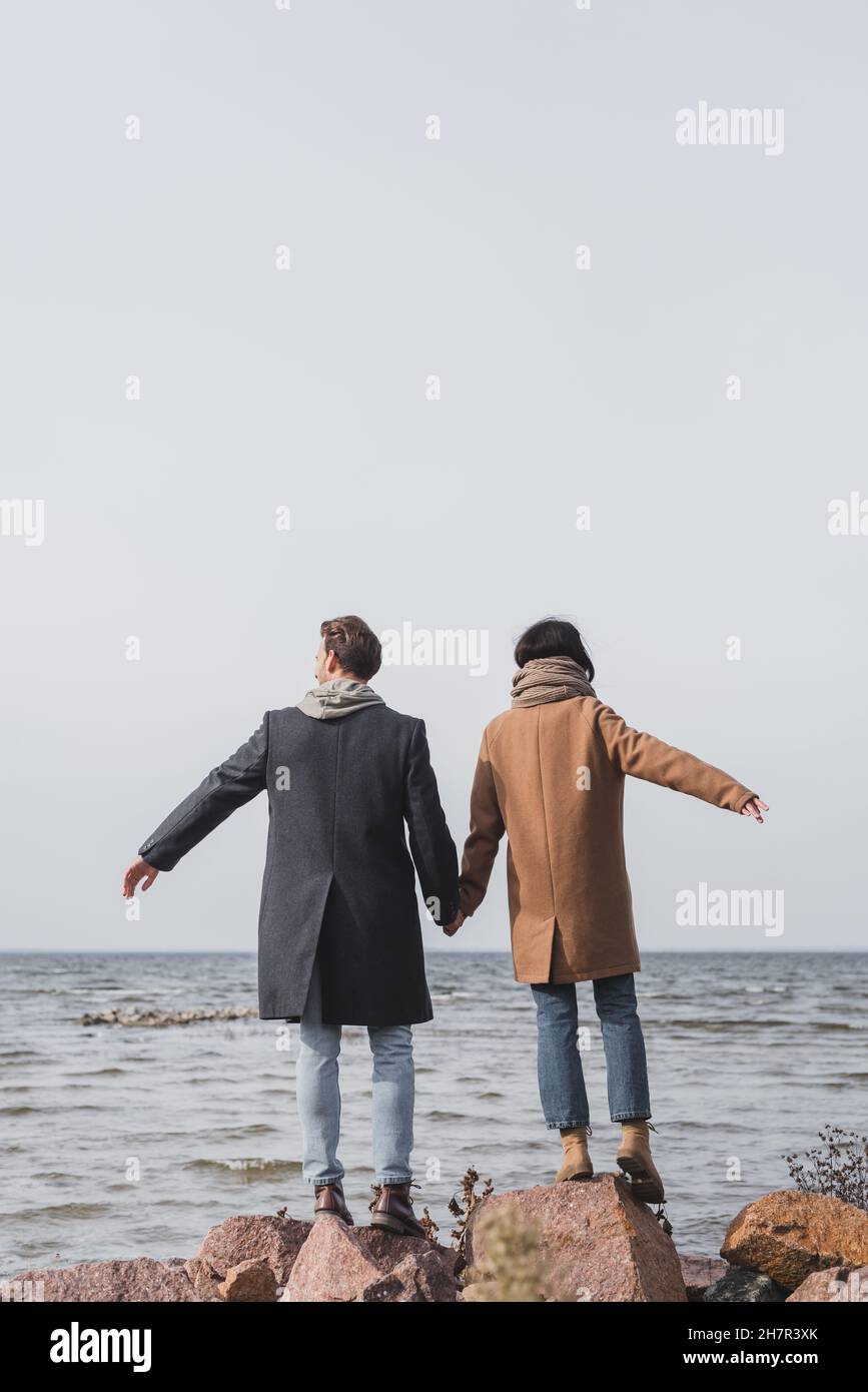 back view of young couple in autumn coats holding hands while standing on sea coast Stock Photo