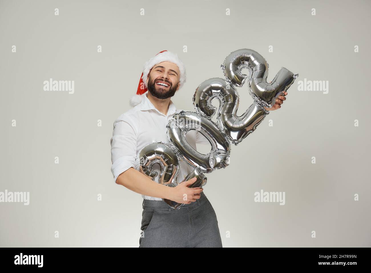 A happy man in a red velvet Santa hat is holding silver balloons in the shape of 2022 like a machine gun. A smiling guy with a beard at a New year par Stock Photo
