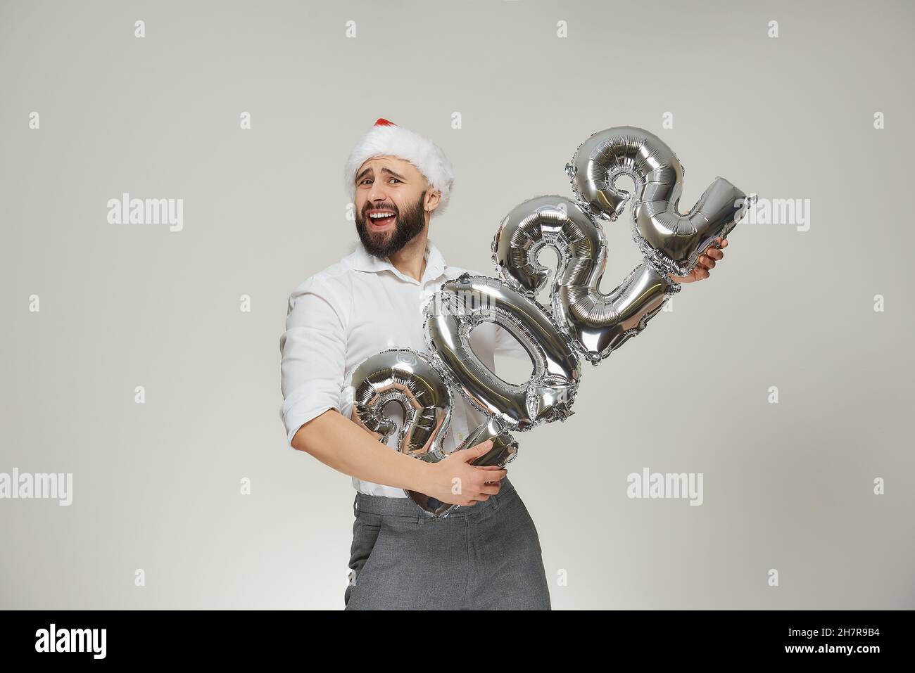 A dashing man in a red velvet Santa hat is holding silver balloons in the shape of 2022 like a machine gun. A smiling guy with a beard at a New year p Stock Photo