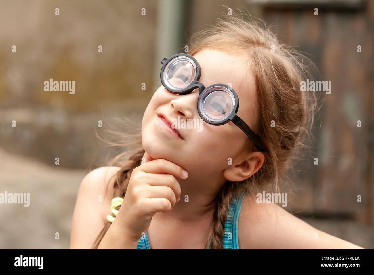 Clever intelligent genius elementary school age girl wearing big funny quirky glasses, wondering, thinking intensely, hand under chin, looking up port Stock Photo