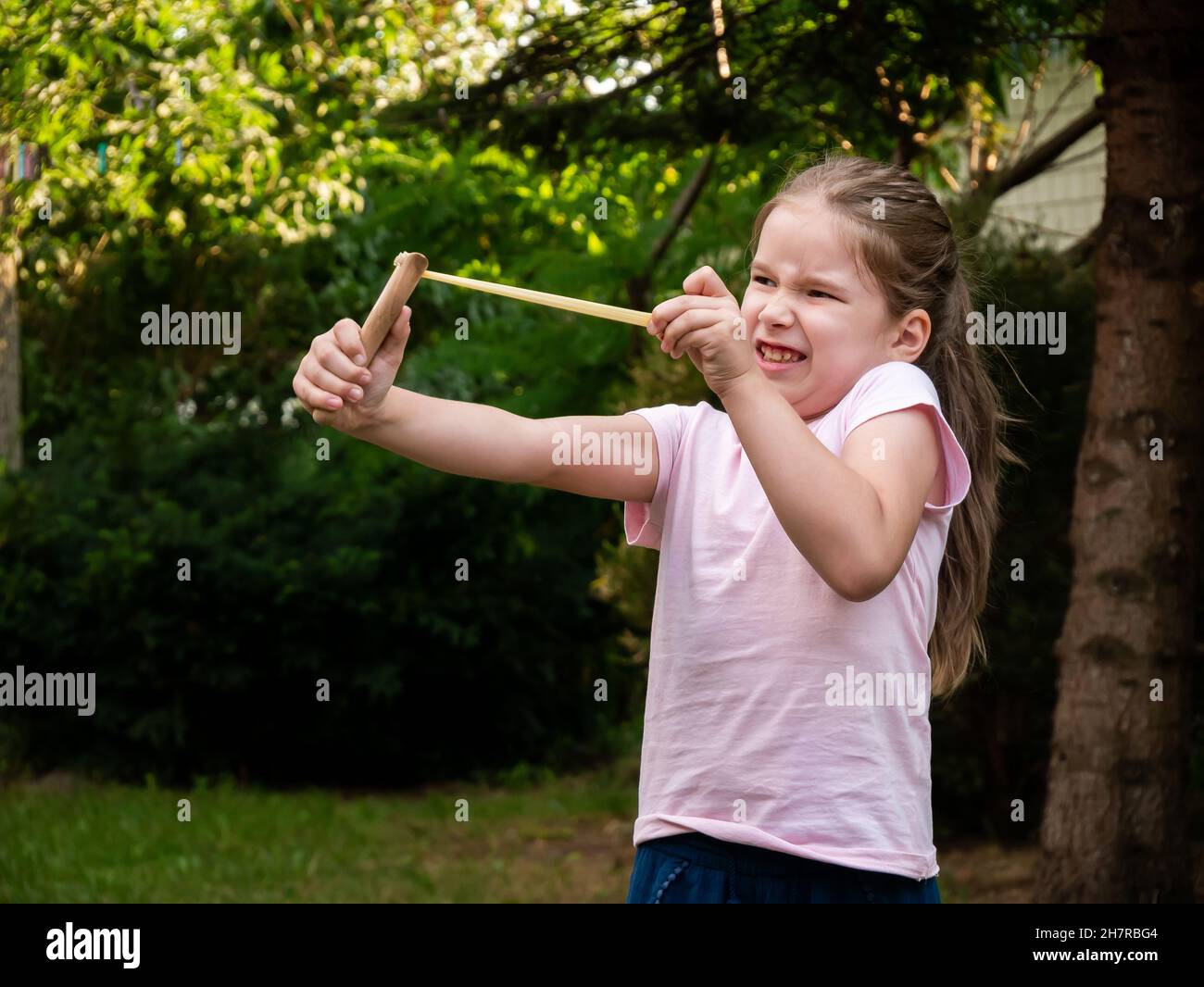 Elementary school age child, young girl pulls back a slingshot shooting, firing a rock making a funny face, outdoors. Toy weapons, tension and strengt Stock Photo