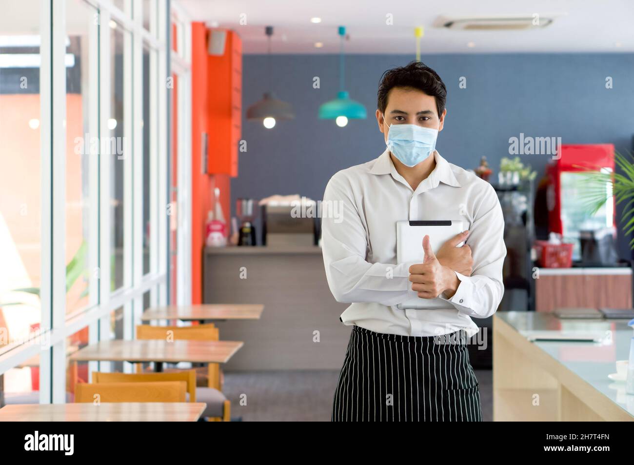 Asian waiter in apron and face mask, raise finger thumb up while holding tablet computer. Stock Photo