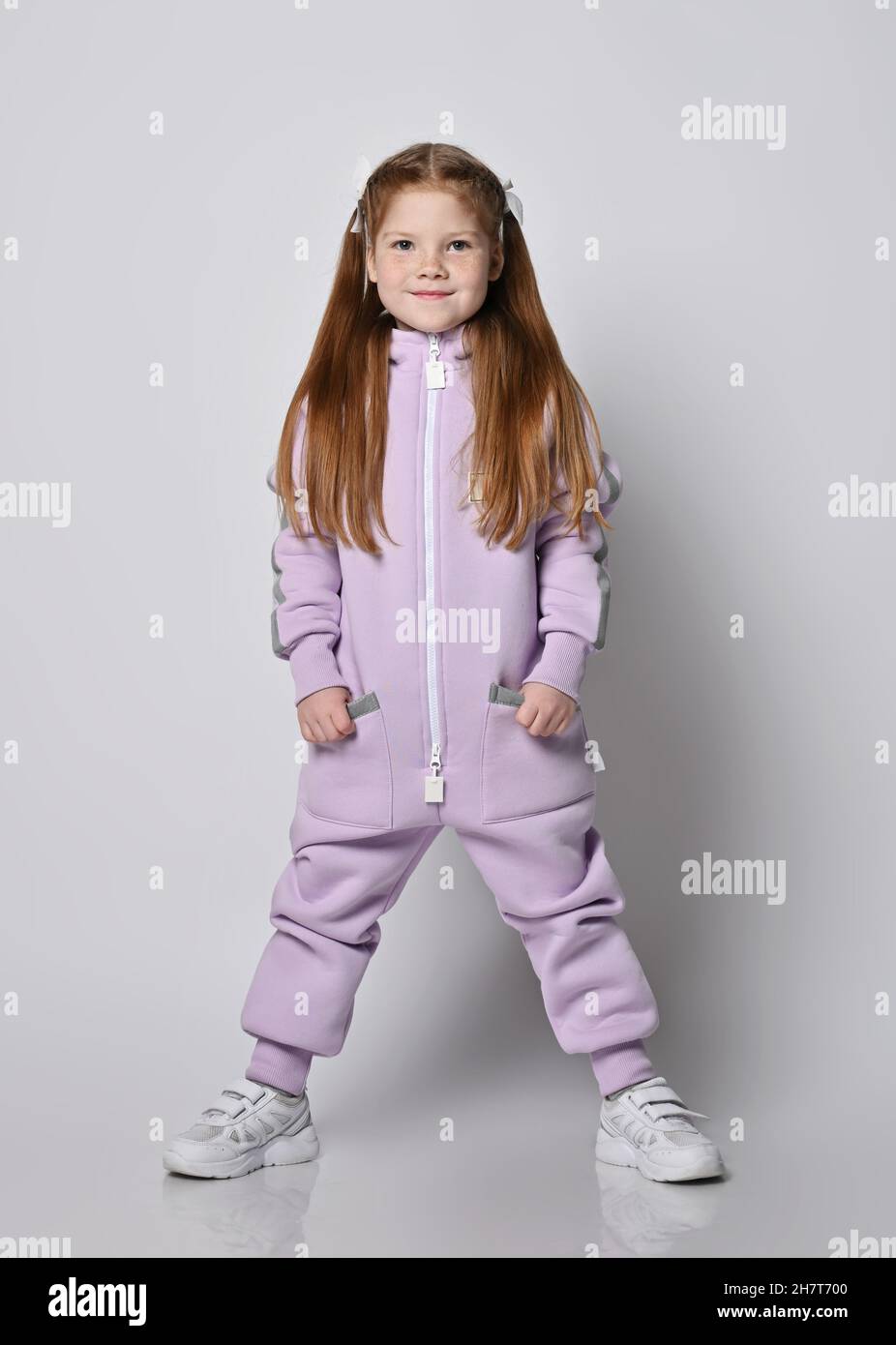 Smiling red-haired kid girl in pink jumpsuit with zipper stands with legs wide apart holding hands in pockets Stock Photo