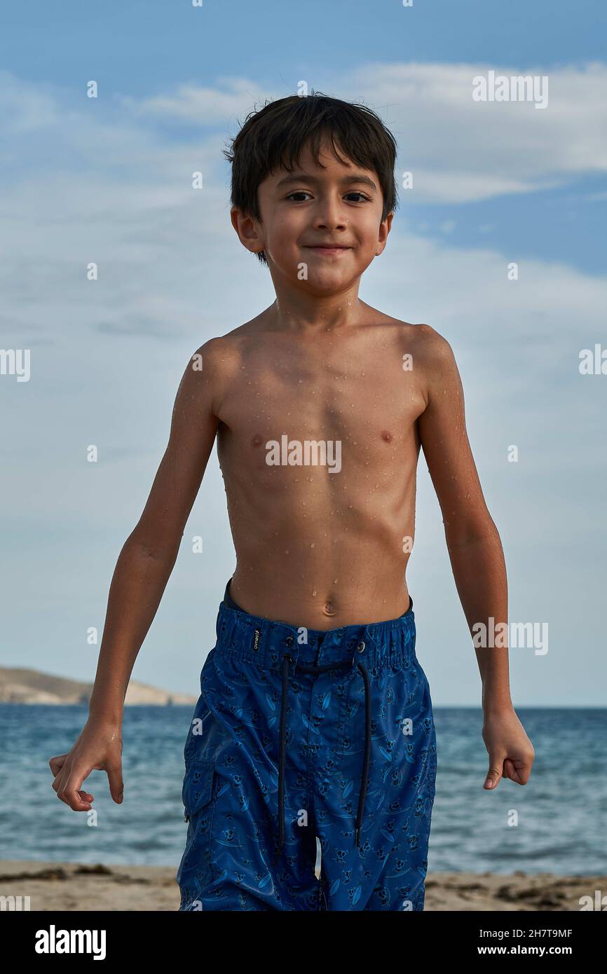 Vertical shot of a little boy with blue swim shorts coming out of a sea with a background of