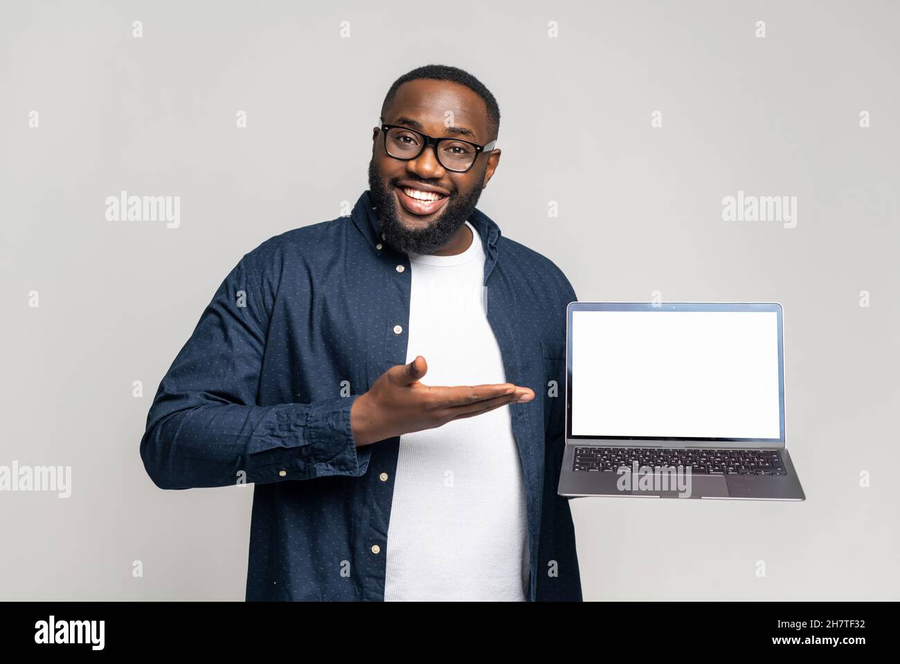 Smiling young African-American guy in casual wear showing laptop with empty display on grey background. Black guy points with palm at blank screen, advertising new app, presentation of newelty Stock Photo
