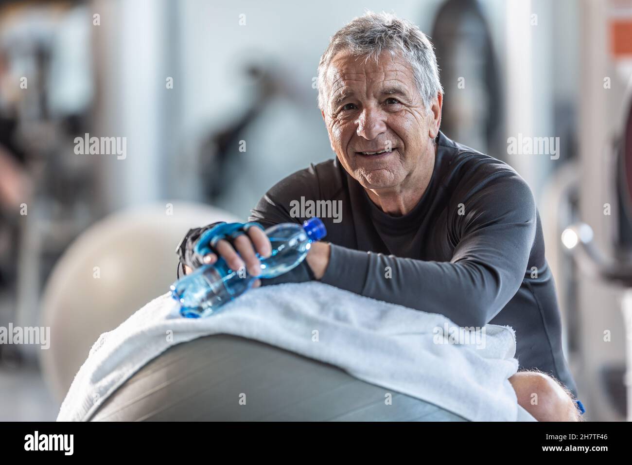 Elderly man resting on an exercise ball in the gym holds a water bottle ...