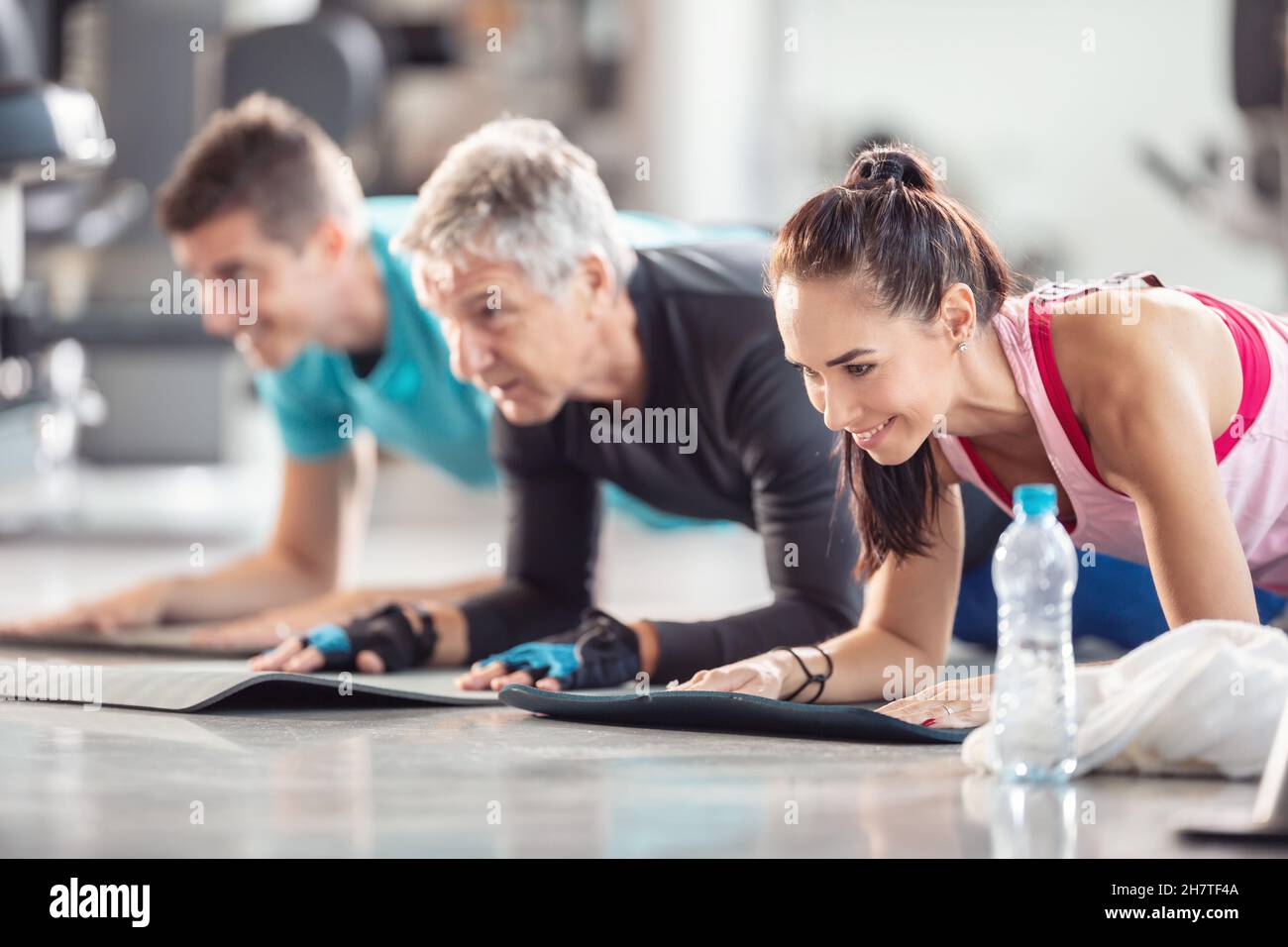 Diverse age group doing elbow planks on mats inside the gym with a plastic water bottle in the front. Stock Photo