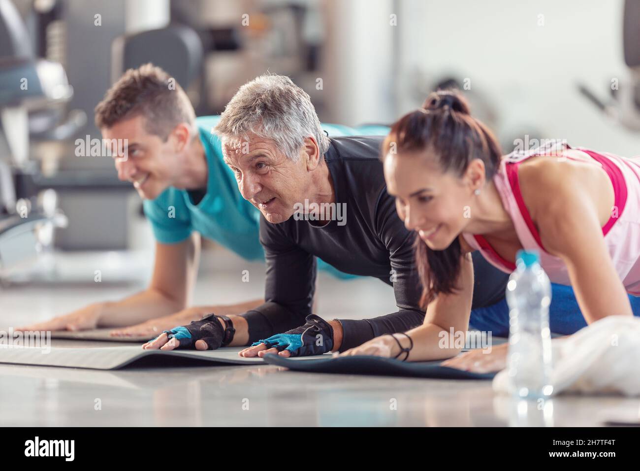 Group of people in various age and gender having fun while doing group exercise of elbow plank in the gym. Stock Photo