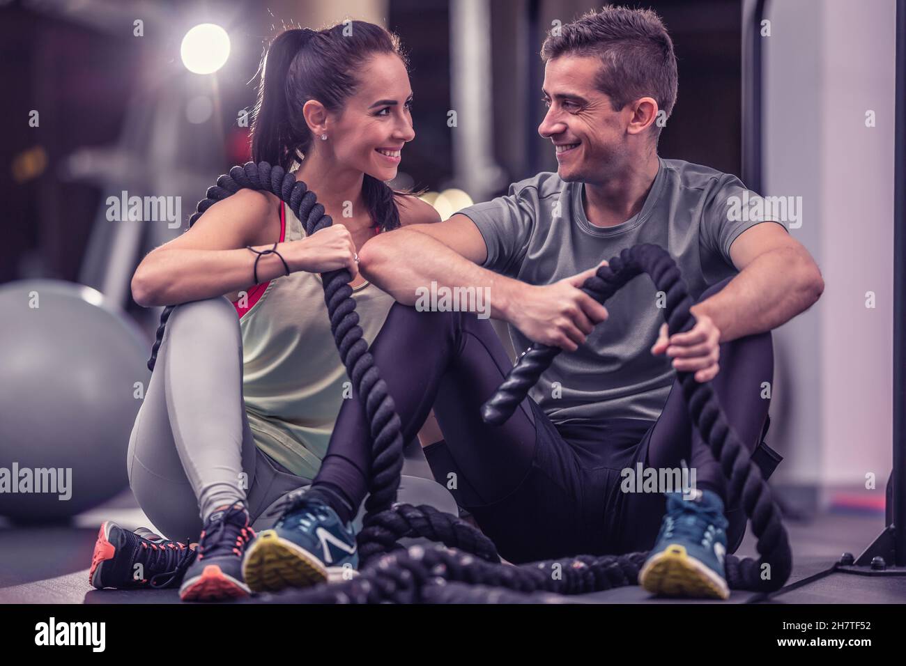A couple that finished battle ropes workout sit together on a ground inside the gym, looking at each other, smiling. Stock Photo