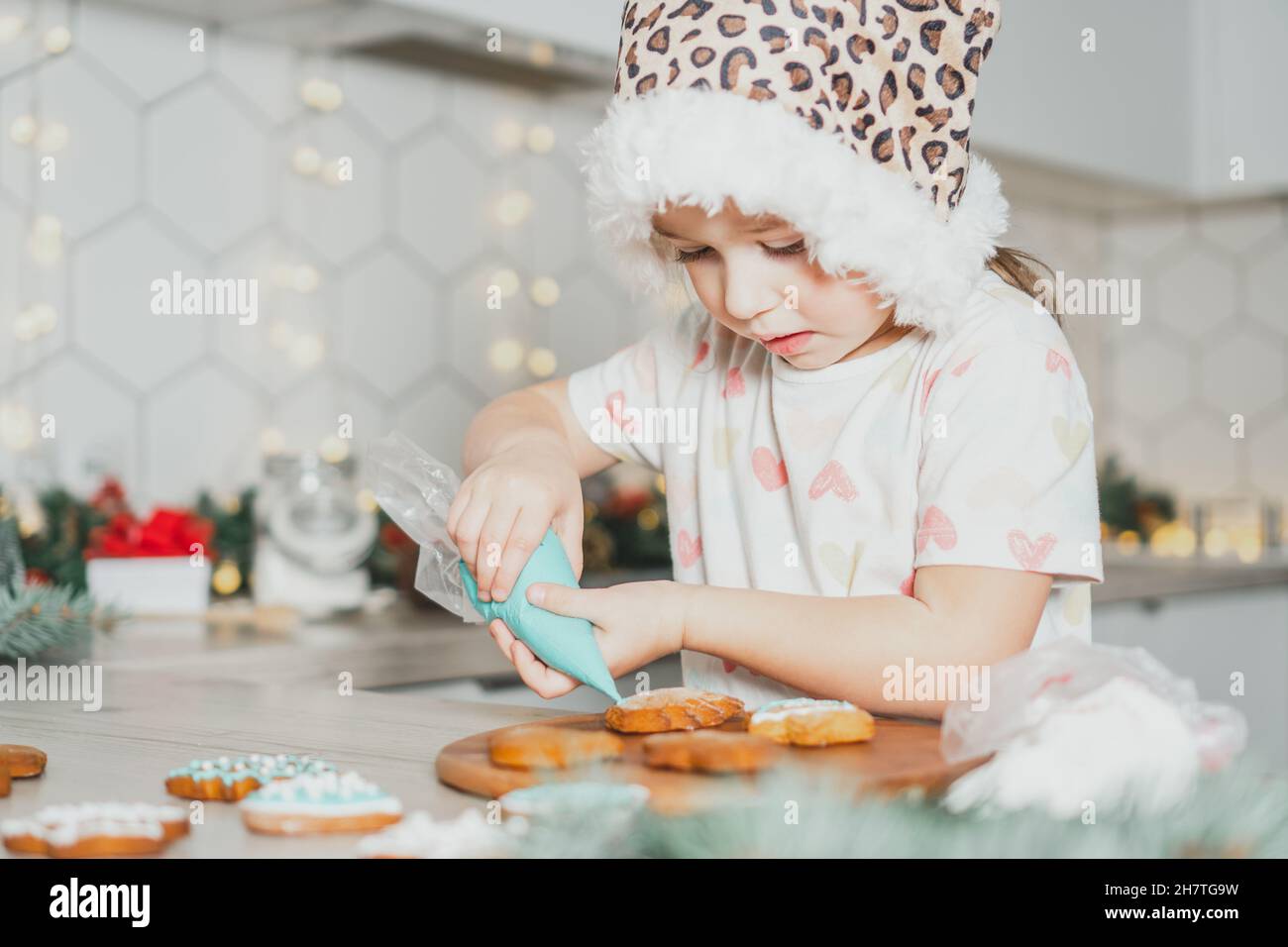 Little dark-haired girl 3 years old in Santa spotted cap decorate gingerbread cookies with icing in white Christmas decorated kitchen. Merry Christmas Stock Photo
